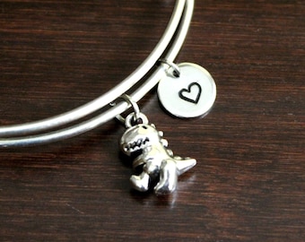 May include: A silver bangle bracelet with a small dinosaur charm and a heart charm. The bracelet is on a dark brown wooden surface.