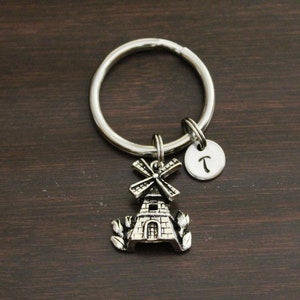 May include: Silver keychain with a windmill charm and a letter 'T' charm. The keychain is on a brown wooden surface.