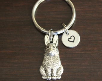 May include: Silver-toned keychain featuring a detailed rabbit charm and a small round tag with a heart outline. The keychain is attached to a silver ring. The background is a dark brown wooden surface.
