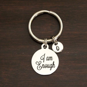 May include: Silver-toned keychain with a large round charm engraved with the words "I am Enough" in elegant script. Includes a smaller charm with the letter "O". The keychain is on a dark brown wooden surface.
