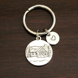 May include: Silver keychain with a charm featuring a small house with a chimney and trees. The charm is attached to a silver key ring with a small heart charm.