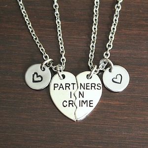 May include: Two silver-toned metal necklaces featuring a split heart pendant engraved with the text PARTNERS IN CRIME. Each necklace includes a small circular charm stamped with a heart symbol, suspended from a matching silver-toned link chain.