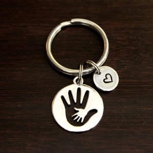 May include: Silver keychain with a circle charm featuring an outline of two hands, one large and one small, holding each other. A smaller heart charm is attached to the ring.