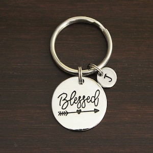 May include: A silver-colored keychain with a large round charm that says "Blessed" in cursive with an arrow and heart design. A smaller charm with the letter "J" hangs next to it. The keychain is on a dark brown wooden surface.