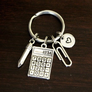 May include: Silver keychain with a calculator, pencil, paperclip, and heart charm. The calculator has the numbers 1234, 789, 456, 123, and 0 on the face.