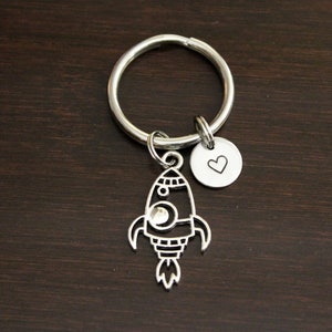 May include: Silver rocket ship keychain with a small heart charm. The rocket ship has a detailed design with a round window and a flame coming out of the back.