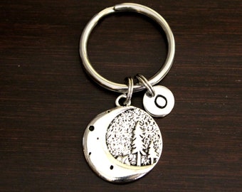 May include: Silver keychain with a crescent moon charm featuring a silhouette of trees and stars. The charm is attached to a silver ring with a small silver disc charm with the letter "O" engraved on it.