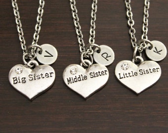 May include: Three silver necklaces with heart-shaped pendants that say "Big Sister", "Middle Sister", and "Little Sister". Each pendant has a small silver circle with a letter on it.