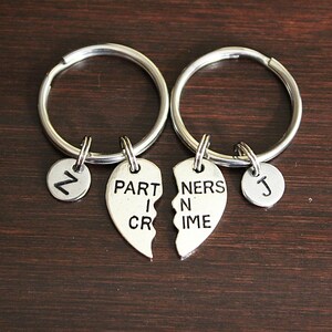 May include: Two silver-toned metal keychains, each featuring a split heart charm. One heart half is stamped with the text PARTNERS IN CRIME, and the other half is stamped with the same text. Each keychain includes a small circular charm with the letter Z or J.