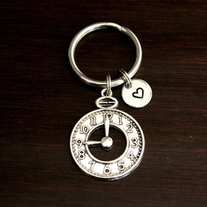 May include: Silver key chain with a clock face charm and a small heart charm. The clock face has Roman numerals and a silver hand.