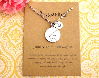 May include: A silver necklace with a round pendant featuring the Aquarius constellation and a small heart charm. The necklace comes on a brown card with the text "Aquarius, January 20 - February 18" and a description of the zodiac sign.