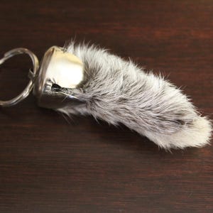 May include: A keychain featuring a silver-colored metal cap and ring, attached to a small, gray and white fur object. The fur has a soft, textured appearance. The item is set against a dark brown wooden surface.