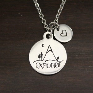 May include: Silver necklace with two charms. The larger charm features a mountain range with trees and the word "EXPLORE". The smaller charm is a heart.