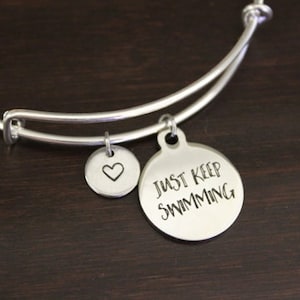 May include: Silver-toned bangle bracelet with two charms. One charm is a small circle with a heart, and the other is a larger circle with the words "JUST KEEP SWIMMING" in a handwritten font. The bracelet is on a dark brown surface.