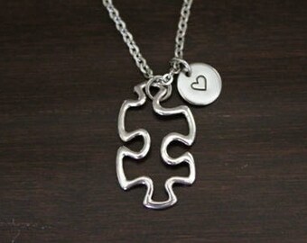 May include: A silver necklace with a puzzle piece pendant and a small heart charm. The necklace is on a dark wooden surface.