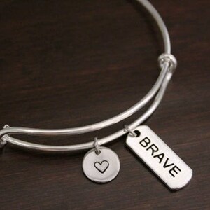 May include: Silver expandable bangle bracelet with two charms. One charm is a round disc with a heart engraved on it. The other charm is a rectangular tag with the word "BRAVE" engraved on it.