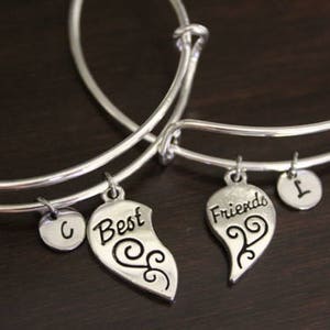 May include: Two silver charm bracelets with heart-shaped charms that say "Best" and "Friends". The charms are connected to the bracelets with silver rings. The bracelets also have silver charms with the letters "C" and "L" on them.