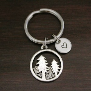 May include: Silver keychain with a round charm featuring two trees inside a circle. A smaller heart charm is attached to the keyring.