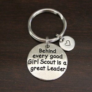 May include: Silver keychain with a round charm that says "Behind every good Girl Scout is a great Leader". A smaller heart-shaped charm is attached to the keyring.