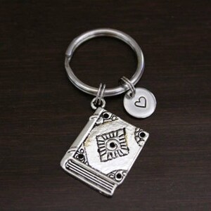 May include: Silver keychain with a book charm and a small heart charm. The book charm has a detailed design and the heart charm has a simple design.