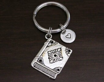 May include: Silver keychain with a book charm and a small heart charm. The book charm has a detailed design and the heart charm has a simple design.
