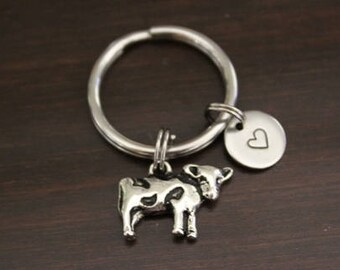May include: Silver cow keychain with a heart charm. The cow is standing on all four legs and has black and white spots. The keychain is attached to a silver key ring.