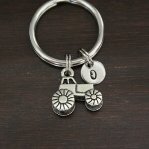 May include: Silver-toned tractor keychain with a round key ring and a small, circular charm with the letter "O". The tractor charm has detailed wheels. The keychain is on a dark brown wooden surface.