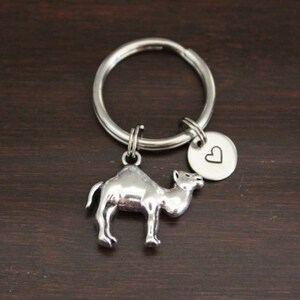 May include: A silver-colored camel keychain with a round charm featuring a heart symbol. The keychain is attached to a silver ring. The background is a dark brown wooden surface.