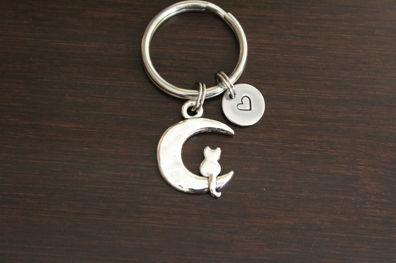 May include: Silver keychain featuring a crescent moon with a cat sitting on it. A small heart charm is attached to the ring.