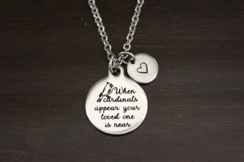 May include: Silver necklace with two charms. The larger charm is round and features a cardinal bird and the text "When cardinals appear your loved one is near". The smaller charm is a heart.