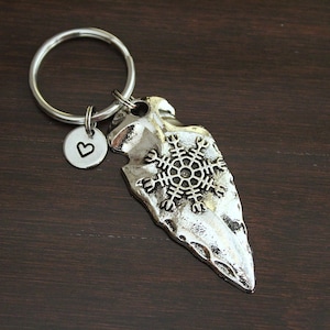 May include: A silver-toned metal keychain featuring an arrowhead-shaped pendant embossed with a detailed Viking-style compass symbol. Attached to the split ring is a small, round silver charm stamped with a black heart icon.