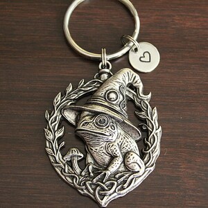 May include: A silver-toned metal keychain featuring a circular pendant with a frog wearing a pointed hat, surrounded by a leafy wreath and a small mushroom. A small round charm with a stamped heart symbol is attached to the split ring.