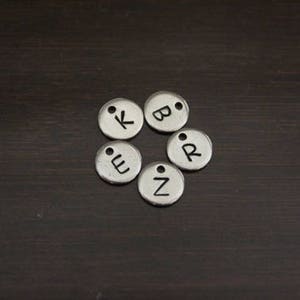 May include: Six silver metal charms with letters K, B, E, Z, and R on a dark brown wooden surface.
