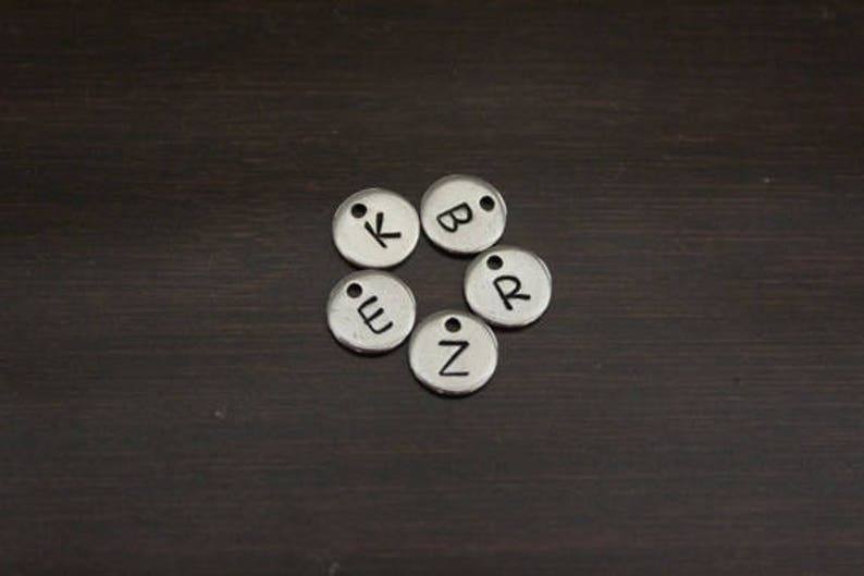 May include: Six silver metal charms with letters K, B, E, Z, and R on a dark brown wooden surface.