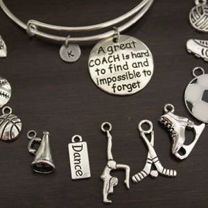 May include: A silver charm bracelet with various sports and dance themed charms. The charms include a basketball, baseball, soccer ball, ice skate, hockey sticks, a megaphone, a dance charm, a gymnast charm, and a charm with the text "A great COACH is hard to find and impossible to forget".