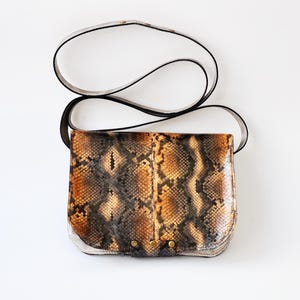 Peut inclure: Un petit sac bandouli&egrave;re en cuir de serpent brun et noir avec une bandouli&egrave;re blanche. Le sac a une fermeture &agrave; rabat et des ferrures dor&eacute;es.