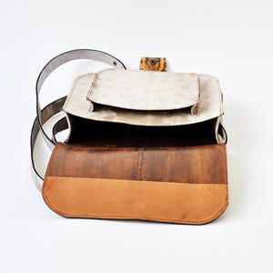 Peut inclure: Un sac &agrave; main en cuir blanc avec un rabat en cuir marron et une petite poche sur le devant. Le sac est ouvert et l'int&eacute;rieur est visible.