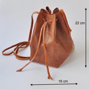 May include: A brown leather drawstring backpack, approximately 16 cm wide and 22 cm tall. The bag has adjustable straps and a drawstring closure. The leather has a textured appearance. The bag is designed to be worn over the shoulders.