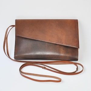 May include: A brown leather crossbody bag with a long, thin strap. The bag has a rectangular shape with a diagonal seam across the front. The leather has a natural, textured appearance.