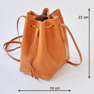 Puede incluir: Bolso tipo cubo de cuero marrón claro con cierre de cordón y correas para el hombro. El bolso mide 22 cm de alto y 16 cm de ancho. El bolso es de cuero liso y flexible.