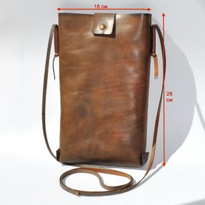 May include: A brown leather crossbody bag, 18 cm wide and 28 cm tall, with a long adjustable strap. The bag features a flap closure with a brass snap. The leather has a distressed finish, giving it a vintage look. The bag is designed to be worn over the shoulder.