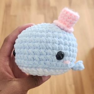 May include: A light blue crocheted whale plushie with a small pink top hat. The whale has black button eyes, a small pink blush, and a white belly. The whale is held in a hand.