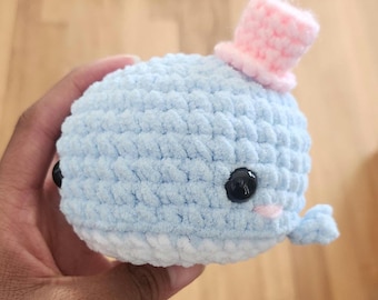 May include: A light blue crocheted whale plushie with a small pink top hat. The whale has black button eyes, a small pink blush, and a white belly. The whale is held in a hand.
