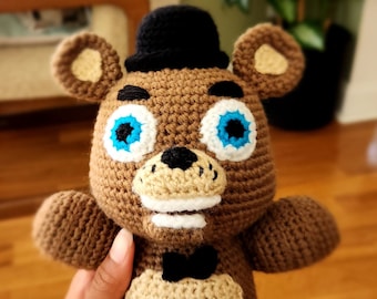 May include: A brown crocheted teddy bear wearing a black top hat and a black bow tie. The bear has blue eyes and a white smile.