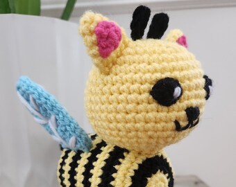 May include: A crocheted bee toy with yellow and black stripes, blue wings, and pink ears. The bee has black eyes and a black nose.
