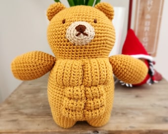 May include: A crocheted teddy bear with a yellow body and brown eyes. The bear is sitting on a wooden table.
