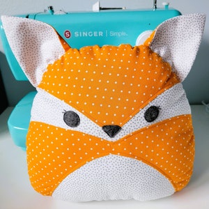 May include: Orange and white fox-shaped pillow with black eyes and a black nose. The pillow is made of fabric with white polka dots on an orange background.