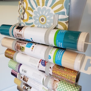 May include: A stack of Cricut brand vinyl rolls in various colours and finishes, including glitter, permanent glossy, and permanent matte. The rolls are labelled with their respective finishes and are organised in a vertical stack.