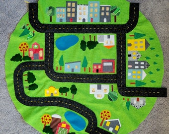 May include: A green felt play mat with a black road design and colorful felt houses, trees, and cars. The mat is perfect for imaginative play and learning.
