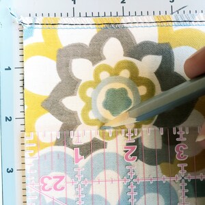 May include: A close-up of a fabric with a floral pattern in shades of yellow, gray, and blue. A ruler with markings in inches is visible, and a pencil is being used to mark a point on the fabric.